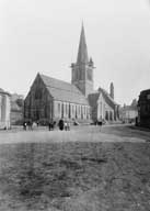 Vue d'ensemble de l'église, côté occidental avec les halles aux bouchers (côté gauche) et l'école primaire des filles (côté droit).- Photographie ancienne, Vannier, 1926-1928.