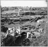 Vue des sites d'exploitation de May-sur-Orne (silos au 1er plan) et de Feuguerolles-sur-Orne (accumulateurs en arrière-plan), et de la passerelle sur l'Orne.- Photographie ancienne, s.d., [1962].